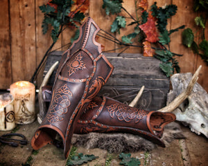 Detailed image of Celtic fantasy greaves with embossed designs, staged in a medieval set with rune-marked candles and animal skulls. The leather is richly dyed and burnished, suitable for Viking warriors, medieval explorers or fantasy LARP characters seeking authentic leg protection.