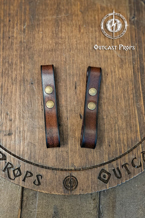 Leather belt pair of clasp mug holder belt strap medieval accessory set of two for LARP cosplay renaissance fair Handcrafted Vegetal Tanned. Two brown leather straps with brass fasteners on a wooden surface with 'Outcast Props' branding.