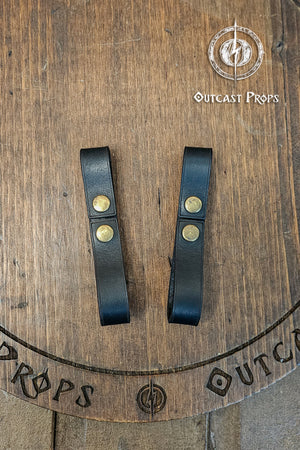 Two black leather straps with gold buttons on a wooden surface, featuring 'Outcast Props' branding. Leather belt pair of clasp mug holder belt strap medieval accessory set of two for LARP cosplay renaissance fair Handcrafted Vegetal Tanned