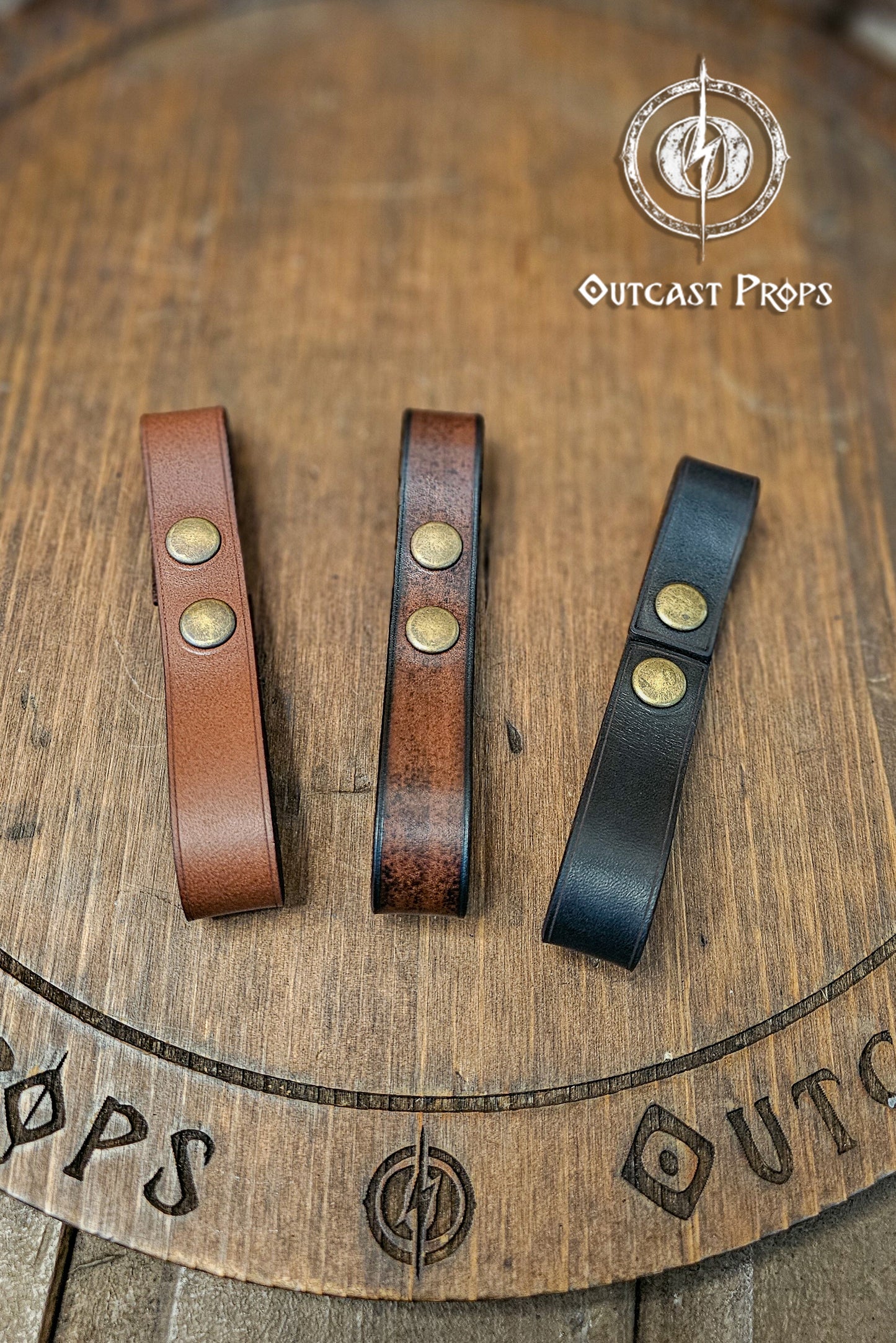 Leather belt pair of clasp mug holder belt strap medieval accessory set of two for LARP cosplay renaissance fair Handcrafted Vegetal Tanned. Three leather straps with metal studs on a wooden surface with 'Outcast Props' branding.