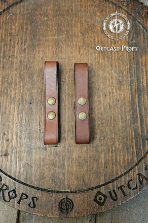 Leather belt pair of clasp mug holder belt strap medieval accessory set of two for LARP cosplay renaissance fair Handcrafted Vegetal Tanned. Two brown leather straps with brass rivets on a wooden surface with 'Outcast Props' branding.