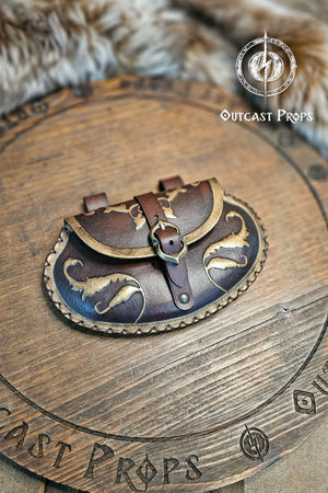 Aged brown renaissance leather belt bag lying flat on engraved wooden board. The small pear shaped pouch shows elegant gold ornaments on flap and edges, with a central brass buckle closure. Two leather loops attach it to a belt. Perfect for LARP, cosplay or renaissance fair noble outfits, this handmade vegetable tanned leather pouch blends style and practicality.