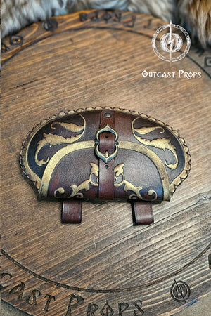 Close-up of pear shape leather belt pouch in aged brown with gold ornaments and metallic buckle, attached to a brown belt. The renaissance bag stands on engraved wooden background with “Outcast Props” logo. It captures historical aesthetics for LARP, cosplay, or renaissance fair noble costumes, made from vegetable tanned leather using eco-friendly dyes.