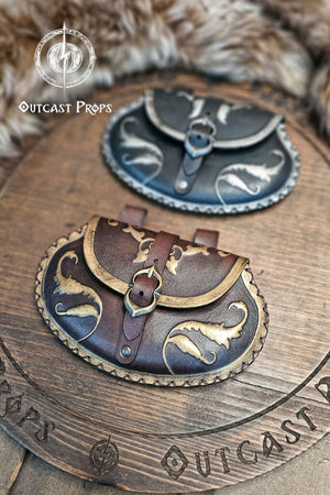 Perspective view showing two noble pear shape leather pouches on wooden board, black with silver and brown with gold. The renaissance belt bags feature detailed painting, scalloped stitching, and brass or nickel buckles. Crafted by Outcast Props for LARP, cosplay, or renaissance fair nobles seeking authenticity and sustainable artisan design.
