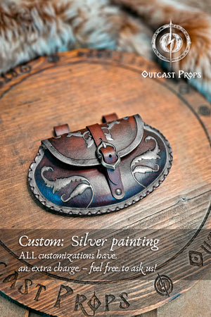 Small pear shape leather pouch in aged black with silver edges and motifs, lying on engraved wooden board with fur background. The renaissance belt bag shows refined silver-painted details, stitched trim, and nickel buckle. Ideal for LARP pouch lovers, medieval fairs, and noble costume accessories handmade from vegetable-tanned leather by Outcast Props.