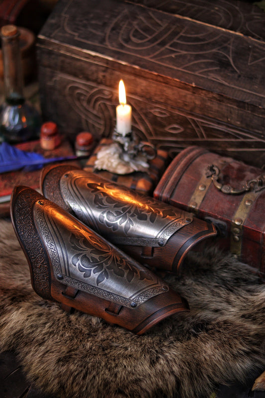 A pair of metal bracers with intricate engravings, placed on a furry surface next to a wooden chest and a candle, possibly for cosplay or theatrical use.