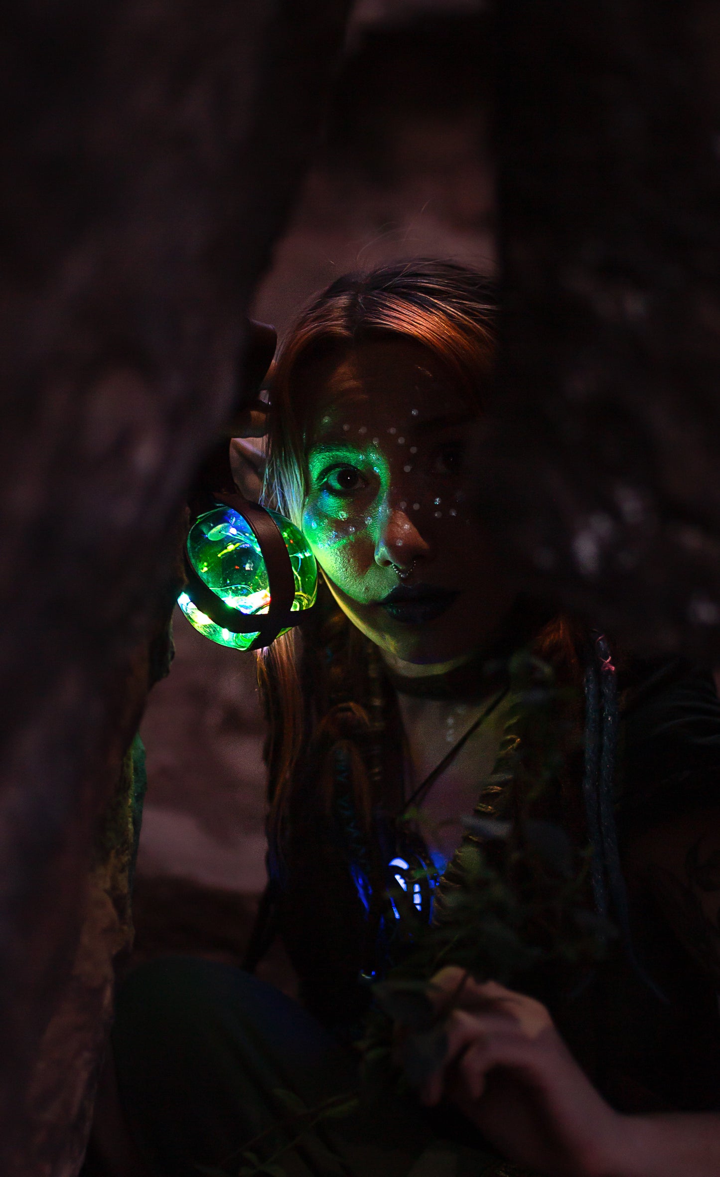 Fantasy character with painted face holding a glowing Magic Light Potion Bottle in a dark, mysterious forest. The orb glows green and multicolored, casting light on her face through leather straps. Perfect accessory for LARP, steampunk, Burning Man, or fantasy cosplay adventures.