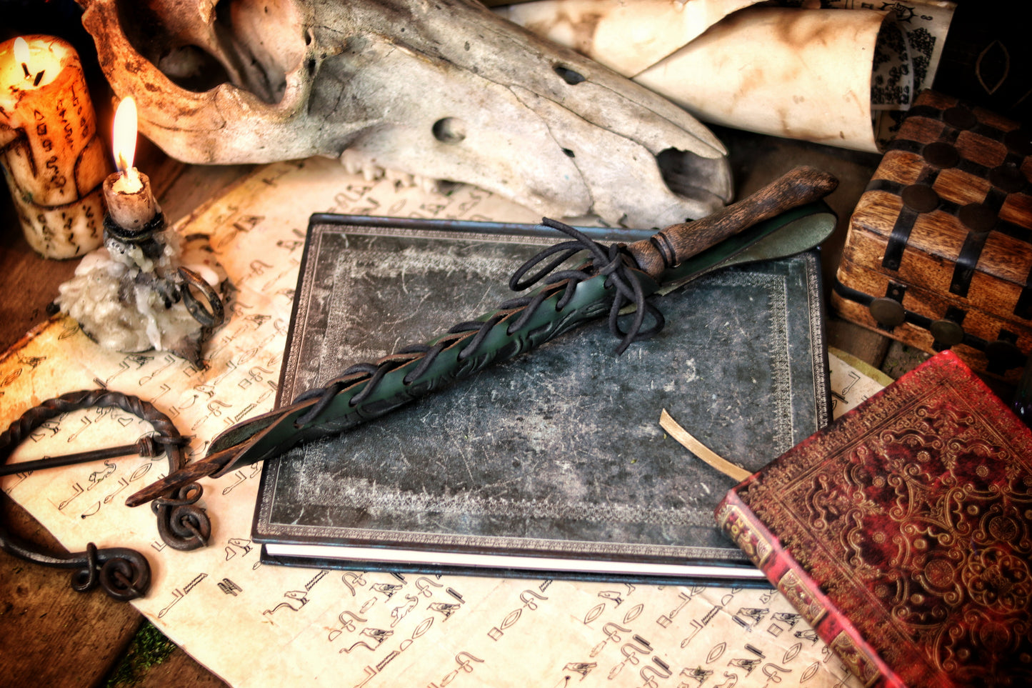 A close-up of an ornate laced green leather wand holster displayed on a table with a decorative backdrop, featuring a leather-bound notebook and a candle. Magic Wand leather Holster Ornated Laced & Aged, for wizard and witch costume belt holster LARP and magic conventions and fair events