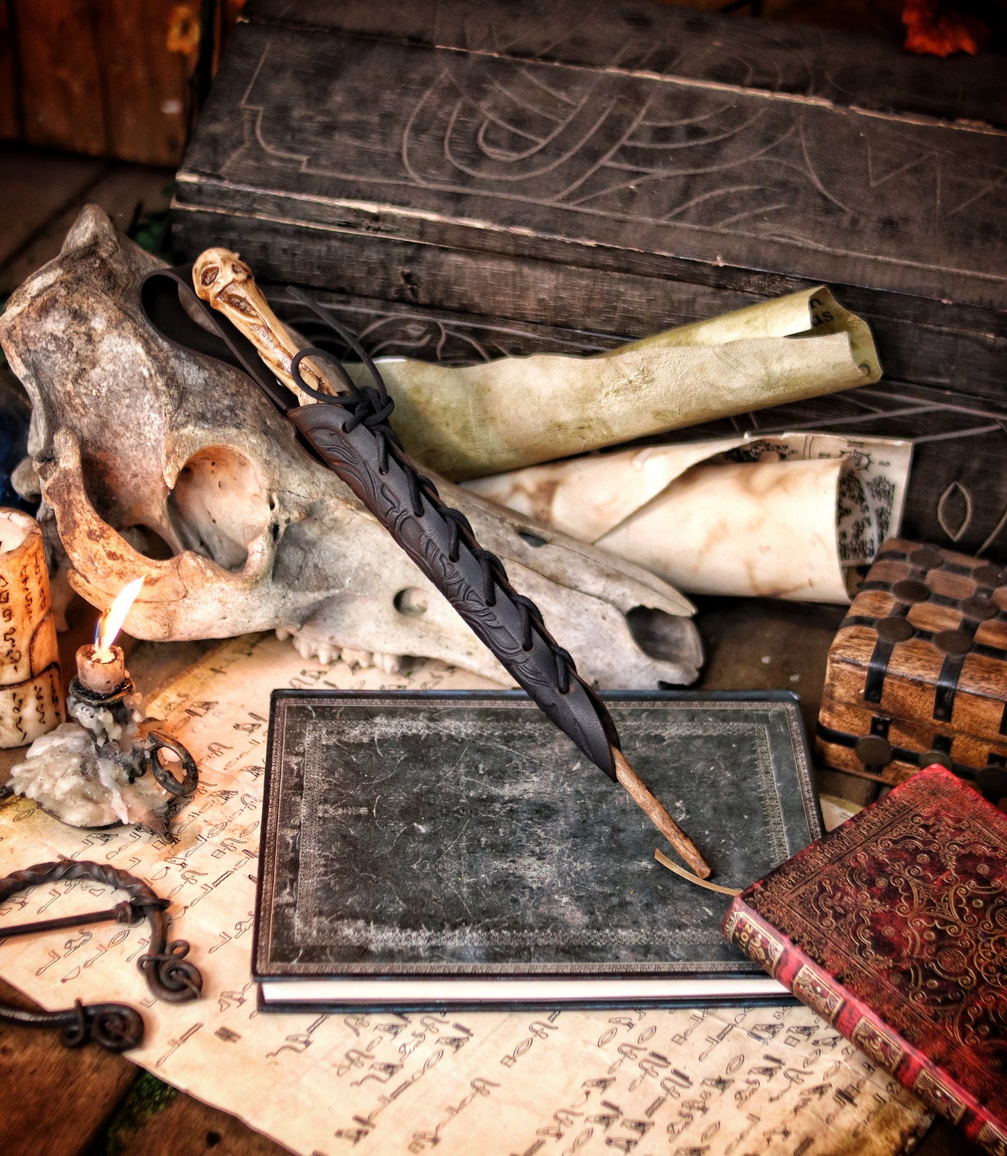 A close-up of an ornate laced black leather wand holster displayed on a table with a decorative backdrop, featuring a leather-bound notebook and a candle. Magic Wand leather Holster Ornated Laced & Aged, for wizard and witch costume belt holster LARP and magic conventions and fair events