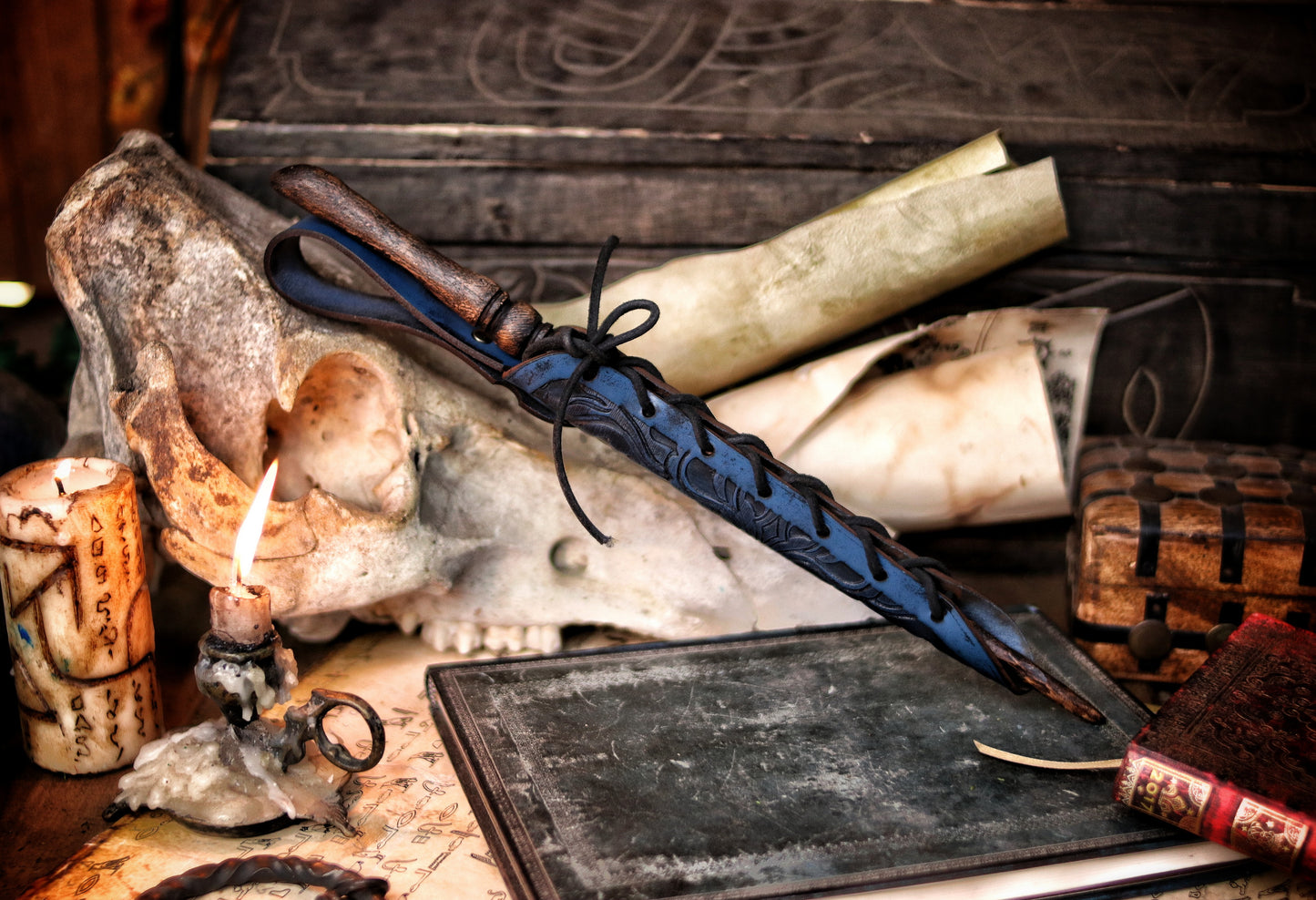 A close-up of an ornate laced blue leather wand holster displayed on a table with a decorative backdrop, featuring a leather-bound notebook and a candle. Magic Wand leather Holster Ornated Laced & Aged, for wizard and witch costume belt holster LARP and magic conventions and fair events