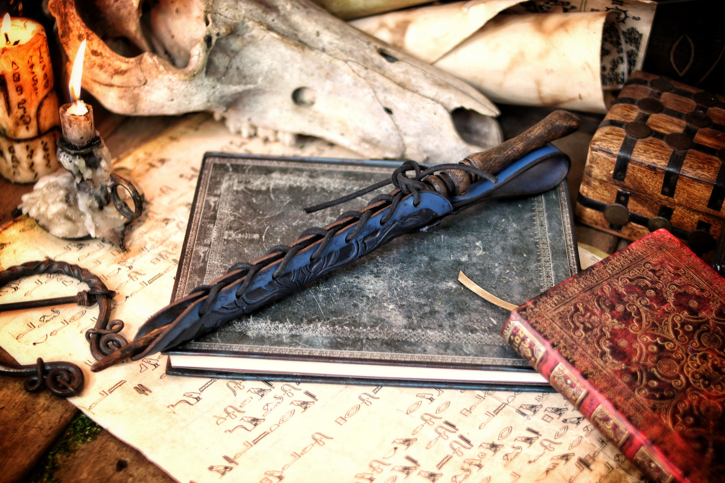 A close-up of an ornate laced blue leather wand holster displayed on a table with a decorative backdrop, featuring a leather-bound notebook and a candle. Magic Wand leather Holster Ornated Laced & Aged, for wizard and witch costume belt holster LARP and magic conventions and fair events