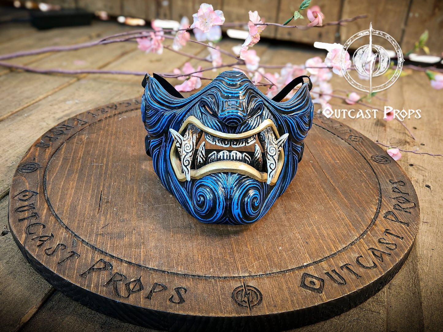 Blue and gold mask on a wooden platform with 'Outcast Props' branding. A detailed and colorful Oni Samurai Mempo mask with intricate hand-painted designs, displayed on a surface with a background of flowers and an old map. Samurai oni mask japanese half oni mask samurai wearable mask for cosplay larp menpo mask scorpion clan mask l5r samurai mask