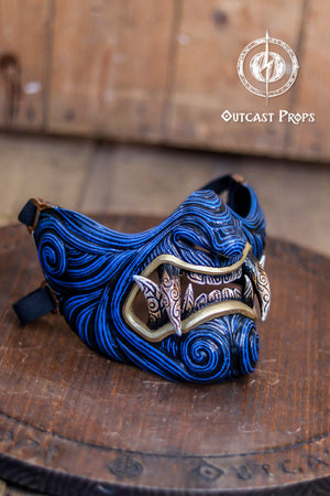 Blue and gold mask on a wooden platform with 'Outcast Props' branding. A detailed and colorful Oni Samurai Mempo mask with intricate hand-painted designs, displayed on a surface with a background of flowers and an old map. Samurai oni mask japanese half oni mask samurai wearable mask for cosplay larp menpo mask scorpion clan mask l5r samurai mask