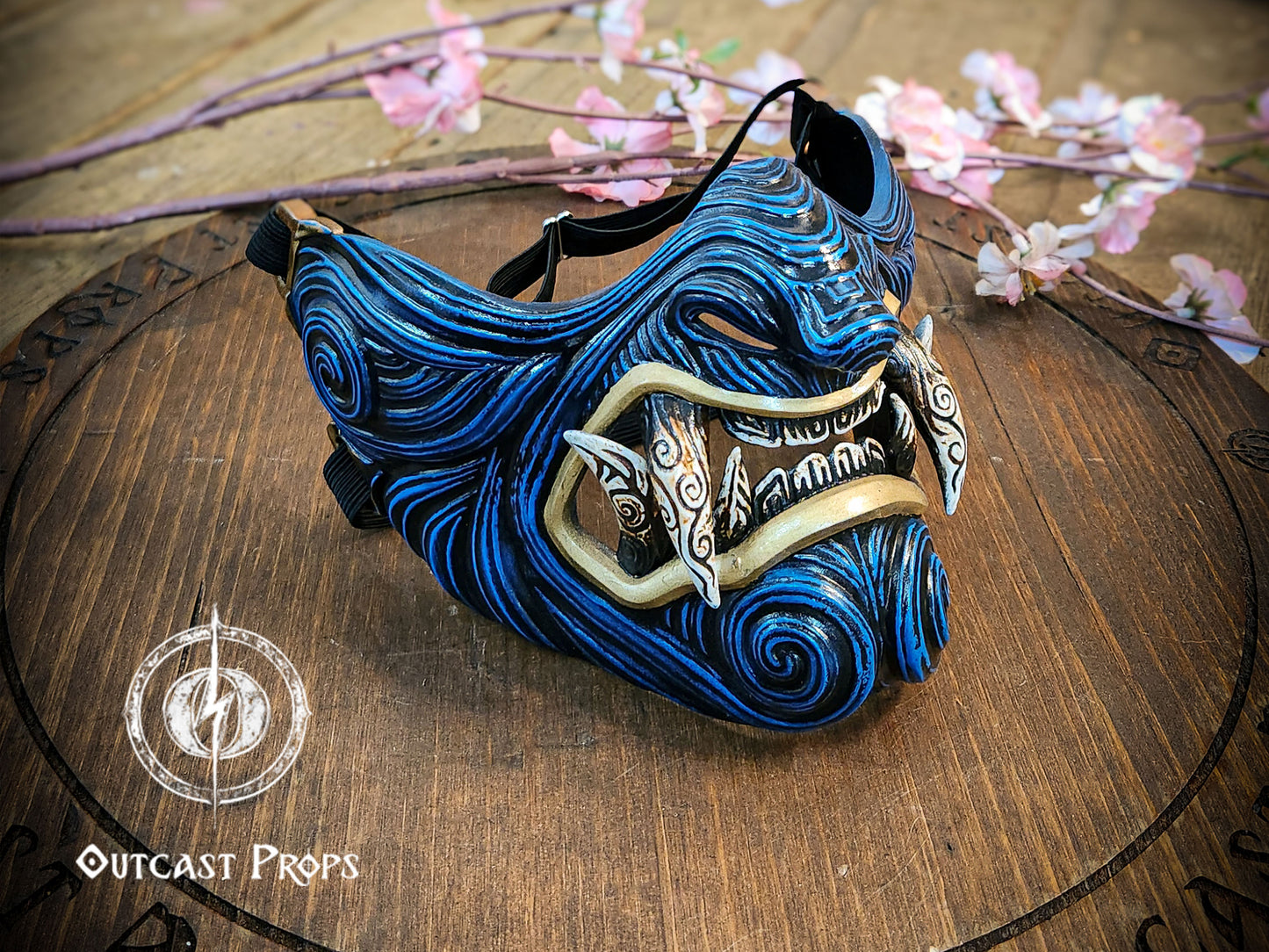 Blue and black decorative mask on a wooden surface with floral decorations. Blue and gold mask on a wooden platform with 'Outcast Props' branding. A detailed and colorful Oni Samurai Mempo mask with intricate hand-painted designs, displayed on a surface with a background of flowers and an old map. Samurai oni mask japanese half oni mask samurai wearable mask for cosplay larp menpo mask scorpion clan mask l5r samurai mask