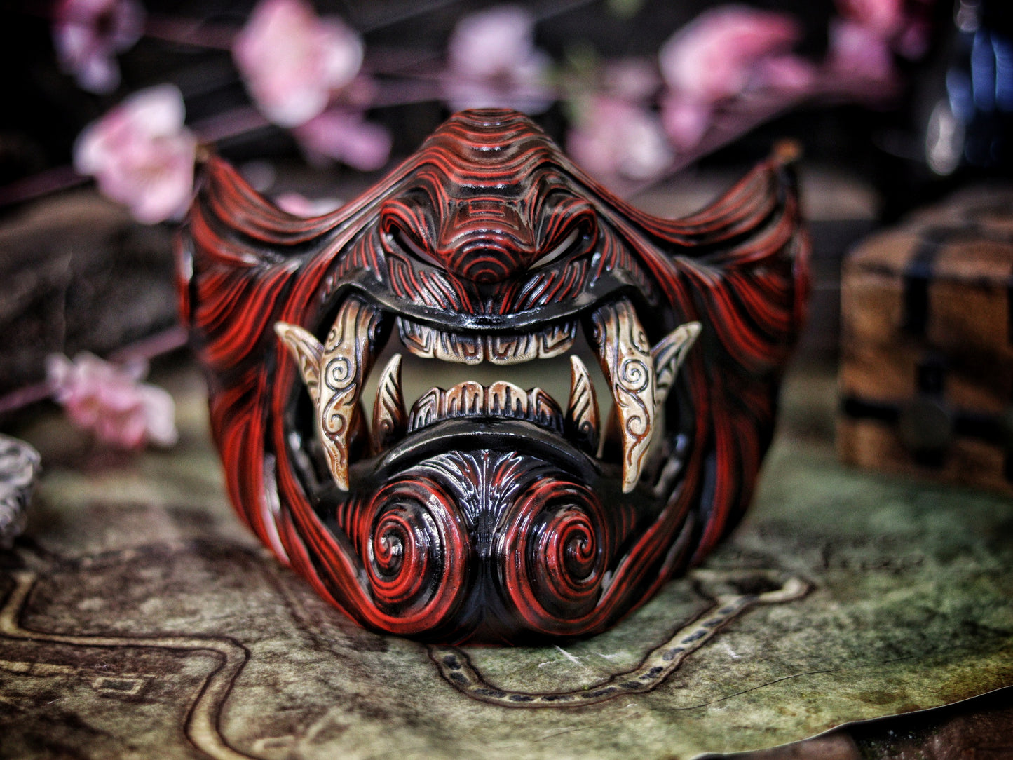 A detailed and colorful Oni Samurai Mempo mask with intricate hand-painted designs, displayed on a surface with a background of flowers and an old map. Samurai oni mask japanese half oni mask samurai wearable mask for cosplay larp menpo mask scorpion clan mask l5r samurai mask