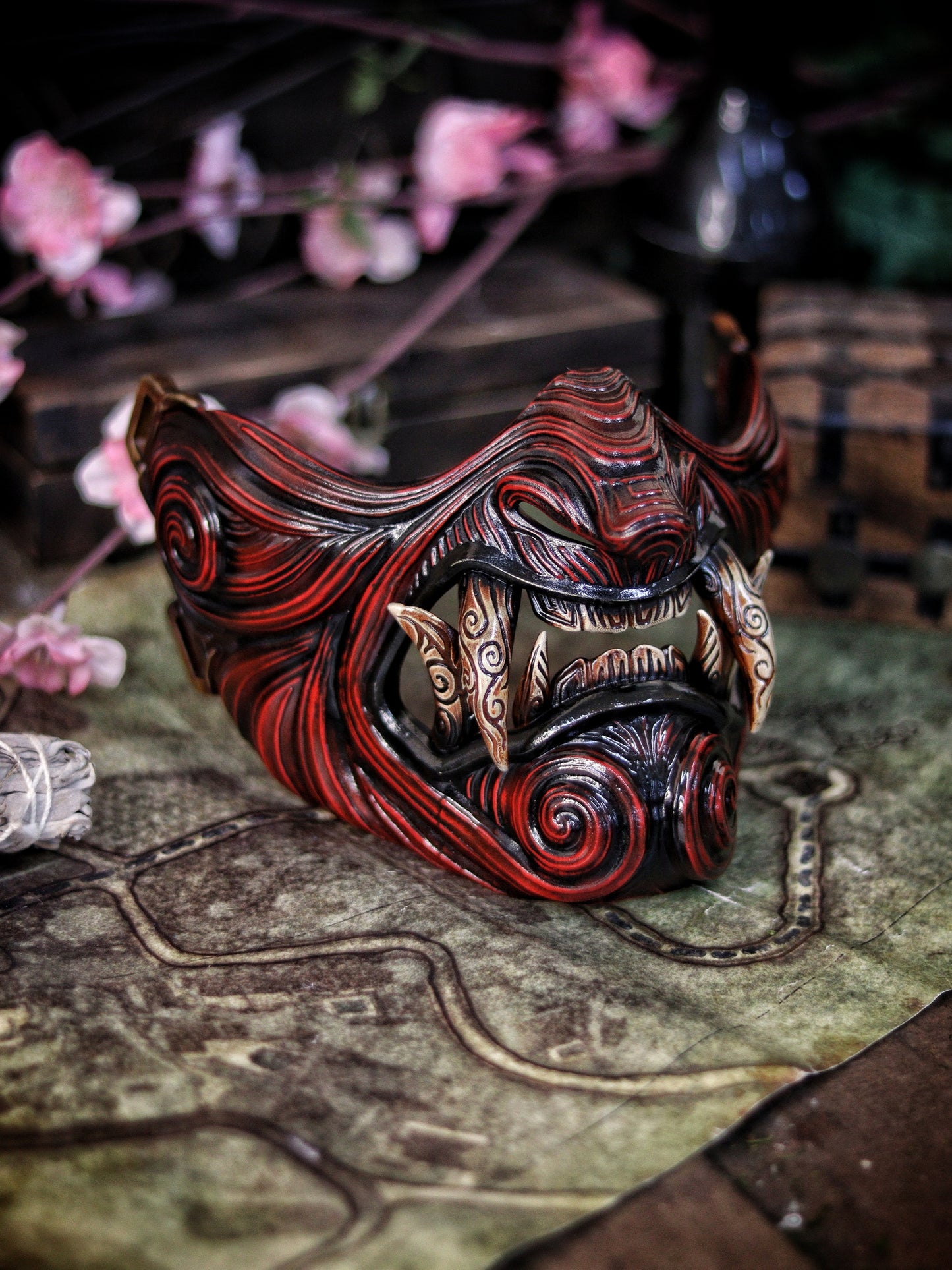 A detailed and colorful Oni Samurai Mempo mask with intricate hand-painted designs, displayed on a surface with a background of flowers and an old map. Samurai oni mask japanese half oni mask samurai wearable mask for cosplay larp menpo mask scorpion clan mask l5r samurai mask