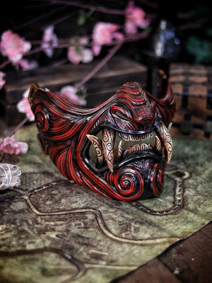 A detailed and colorful Oni Samurai Mempo mask with intricate hand-painted designs, displayed on a surface with a background of flowers and an old map. Samurai oni mask japanese half oni mask samurai wearable mask for cosplay larp menpo mask scorpion clan mask l5r samurai mask