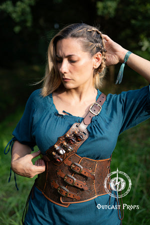 Woman wearing a leather harness in a natural setting with 'Outcast Props' branding. A handmade leather potion holder with ornate embossed patterns, designed to hold five small glass vials. The holder is displayed on a furry surface, and the vials have cork stoppers. Ornated Belt potion holder | 5 potion Belt carrier | LARP, cosplay, fantasy alchemist accessory, adventurers, steampunk, alchemist, healers