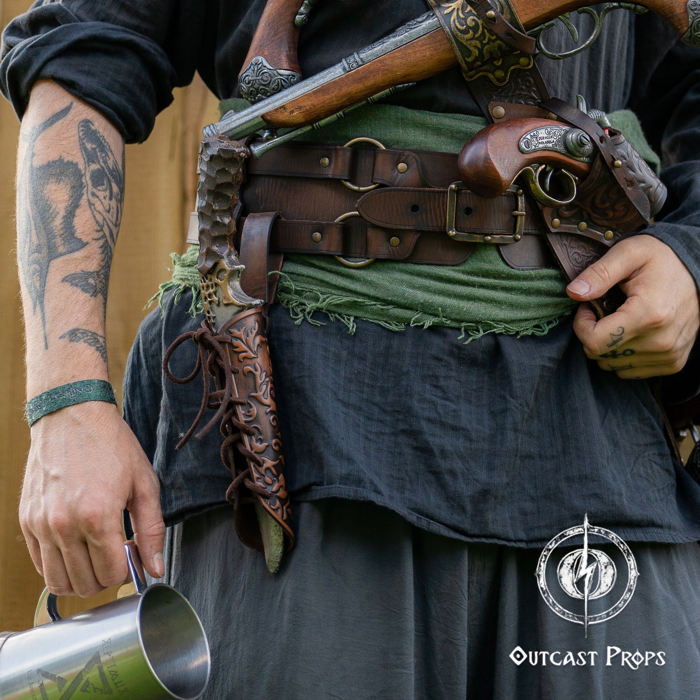Person in historical or fantasy costume with tattoos, holding a cup, and wearing a belt with gear, with 'Outcast Props' logo. Handmade leather dagger sheath | Ornate LARP scabbard | Viking, medieval, fantasy belt holster for daggers | Ren Faire cosplay accessory