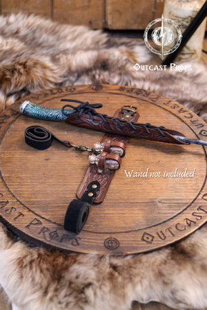 Brown leather wand holster garter displayed on fur background, opened to show all parts: embossed leather panel, thigh strap, bottle holders and lace-up wand sheath. A wooden wand rests inside (not included). Clear product layout for brown leather wizard garter, potion holster for thigh, LARP fantasy leg harness for wands and vials.