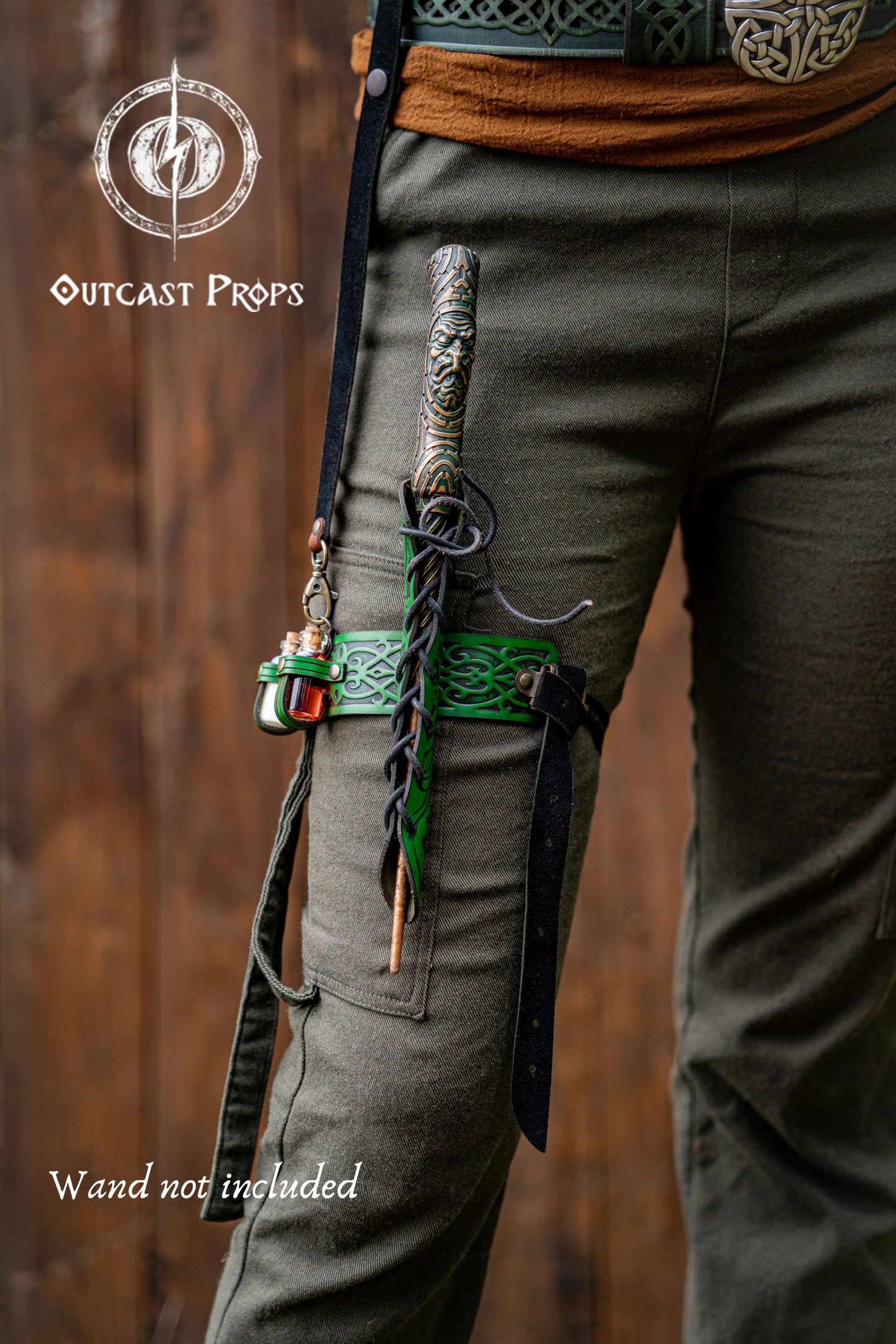 Person wearing green pants with a sword on a wooden background, featuring 'Outcast Props' branding. Cosplayer wears a green embossed leather wand holster garter with potion bottles and a rustic fantasy look. The wand holster wraps around the thigh, secured by adjustable straps. A perfect cosplay wand holder for witches, druids, or mages. The holster doubles as a larp potion holster, keeping tools of magic close. Designed for immersive fantasy costumes and magic users who attend LARP, Ren Faires or wizard