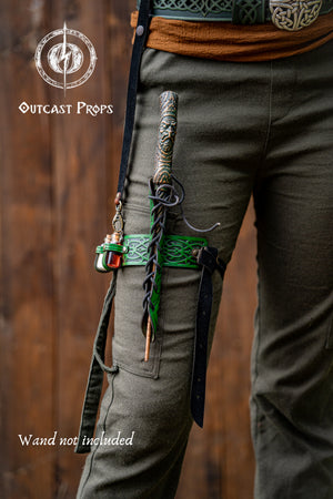 Person wearing green pants with a sword on a wooden background, featuring 'Outcast Props' branding. Cosplayer wears a green embossed leather wand holster garter with potion bottles and a rustic fantasy look. The wand holster wraps around the thigh, secured by adjustable straps. A perfect cosplay wand holder for witches, druids, or mages. The holster doubles as a larp potion holster, keeping tools of magic close. Designed for immersive fantasy costumes and magic users who attend LARP, Ren Faires or wizard