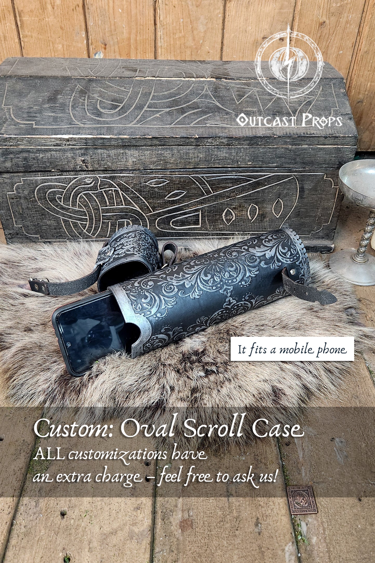 Close-up of a custom oval leather scroll case laying on fur with a medieval chest in the background. The case is hand-tooled in silver over black leather with floral arabesques. A smartphone is partially inserted, showing scale and versatility. This leather scroll holder fits documents, maps, or Magic playmats. Ideal for fantasy cosplay, witch accessories, or Renaissance fair props. Handcrafted by Outcast Props with premium materials and epic painting.