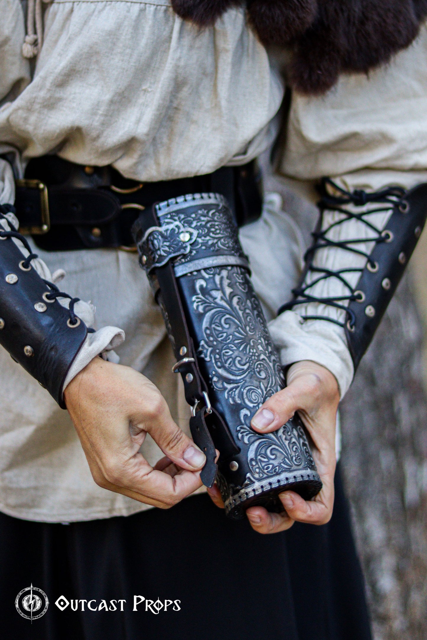 Close-up of a person fastening a black leather scroll case with ornate silver engravings, wearing matching bracers and medieval shirt. Perfect for LARP, fantasy events or Renaissance fairs. Historical leather accessory for scribes, magicians or noble characters. Handcrafted with premium vegetable-tanned leather and embossed in baroque floral pattern. Ideal for medieval.Ornated Leather Scroll Case: Exquisite document tube for cosplay and LARP, Messengers, Magicians, steampunk, reanaissance, fantasy costumes.