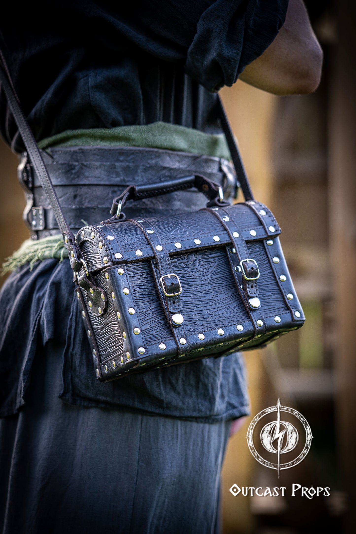 Person holding a black leather bag with studs outdoors, featuring 'Outcast Props' logo. Person wearing a dark outfit with a leather bag, holding a belt with multiple buckles. Man wearing a pirate chest leather bag with studs, standing outdoors. Leather treasure chest pirate chest bag larp pirate backpack treasure chest purse pirate accessories cosplay renfaire pouch belt bag