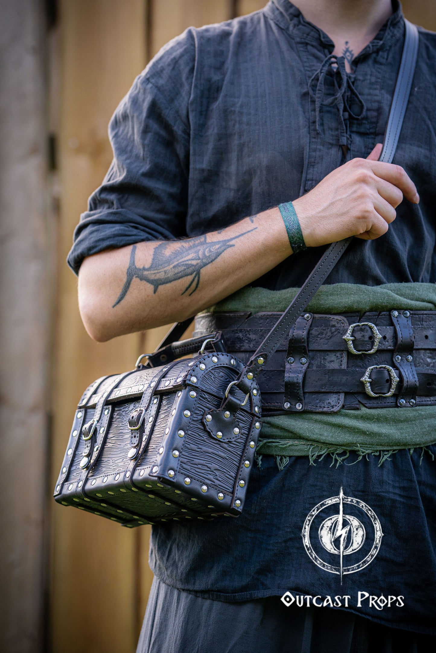 Person wearing a dark outfit with a leather bag, holding a belt with multiple buckles. Man wearing a pirate chest leather bag with studs, standing outdoors. Leather treasure chest pirate chest bag larp pirate backpack treasure chest purse pirate accessories cosplay renfaire pouch belt bag