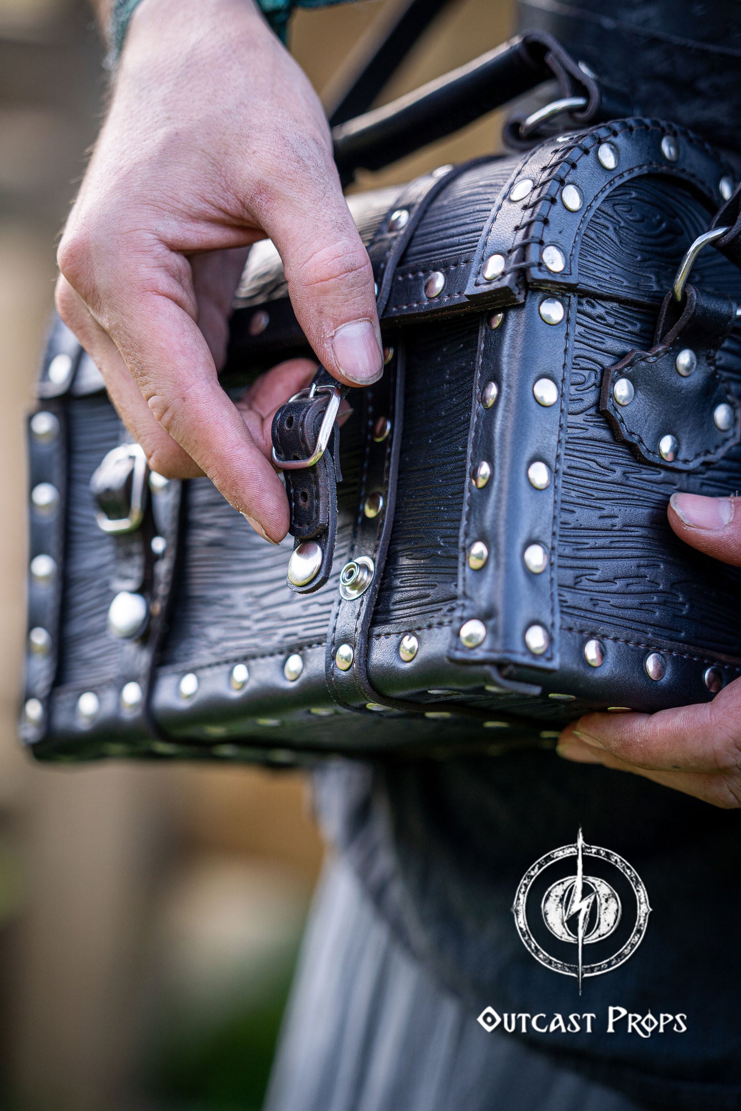 Person holding a black leather bag with studs outdoors, featuring 'Outcast Props' logo. Person wearing a dark outfit with a leather bag, holding a belt with multiple buckles. Man wearing a pirate chest leather bag with studs, standing outdoors. Leather treasure chest pirate chest bag larp pirate backpack treasure chest purse pirate accessories cosplay renfaire pouch belt bag