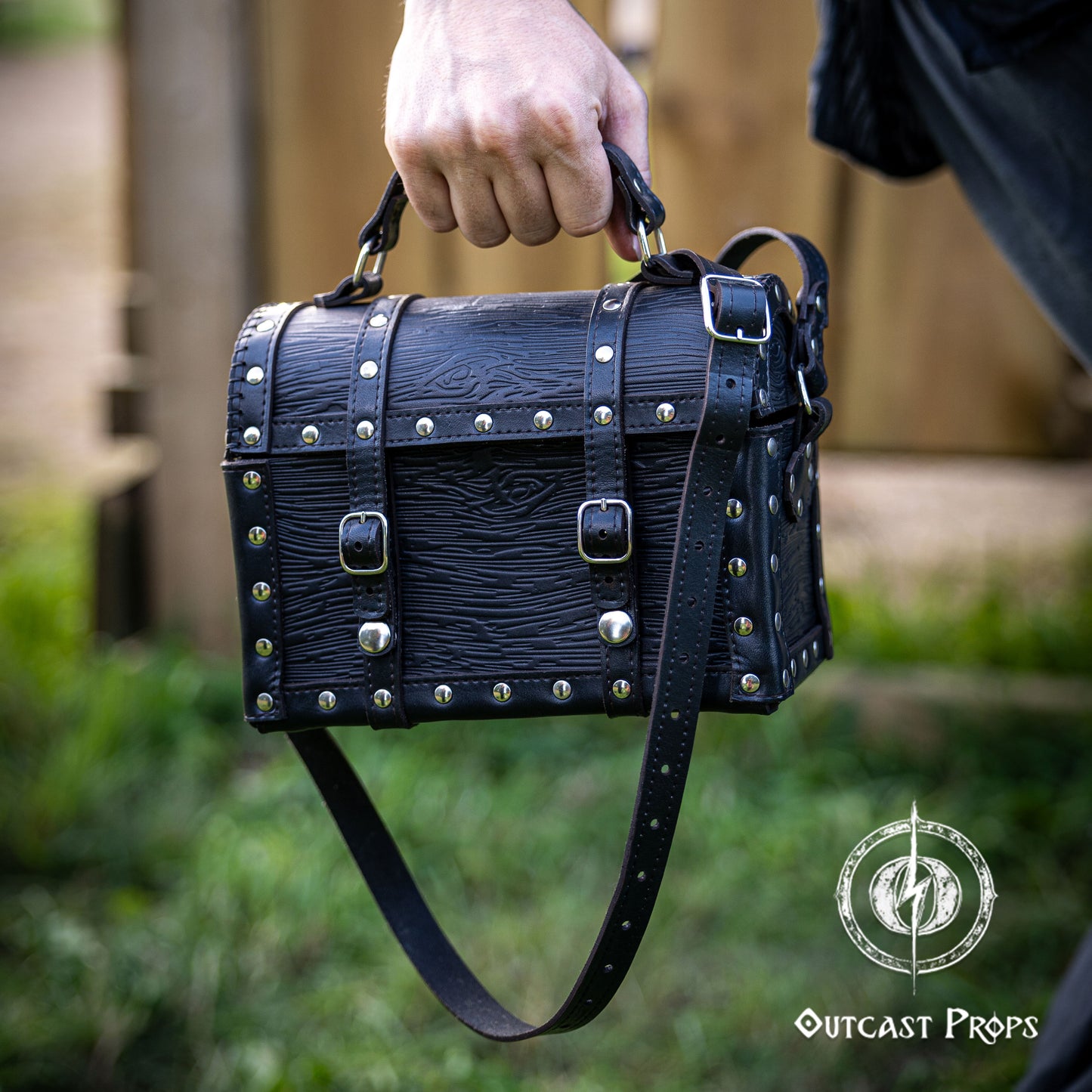 Black leather bag with studs held by a hand outdoors, featuring 'Outcast Props' logo. Person holding a black leather bag with studs outdoors, featuring 'Outcast Props' logo. Person wearing a dark outfit with a leather bag, holding a belt with multiple buckles. Man wearing a pirate chest leather bag with studs, standing outdoors. Leather treasure chest pirate chest bag larp pirate backpack treasure chest purse pirate accessories cosplay renfaire pouch belt bag