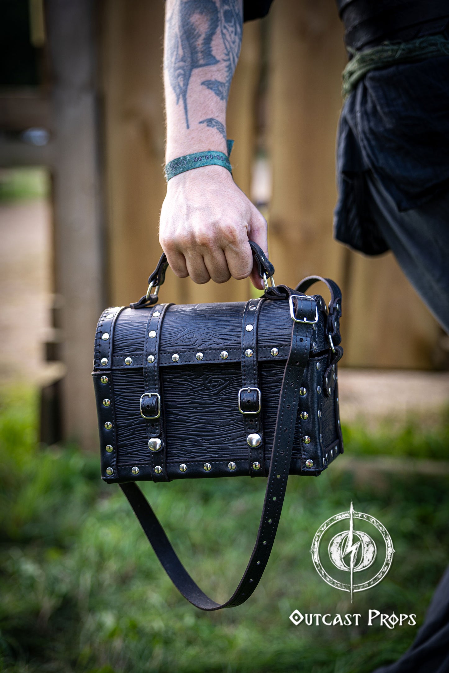 Person holding a black leather bag with studs outdoors, featuring 'Outcast Props' logo. Person wearing a dark outfit with a leather bag, holding a belt with multiple buckles. Man wearing a pirate chest leather bag with studs, standing outdoors. Leather treasure chest pirate chest bag larp pirate backpack treasure chest purse pirate accessories cosplay renfaire pouch belt bag