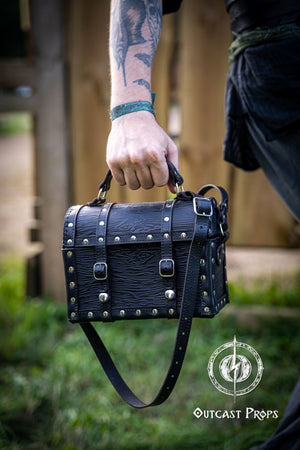 Person holding a black leather bag with studs outdoors, featuring 'Outcast Props' logo. Person wearing a dark outfit with a leather bag, holding a belt with multiple buckles. Man wearing a pirate chest leather bag with studs, standing outdoors. Leather treasure chest pirate chest bag larp pirate backpack treasure chest purse pirate accessories cosplay renfaire pouch belt bag