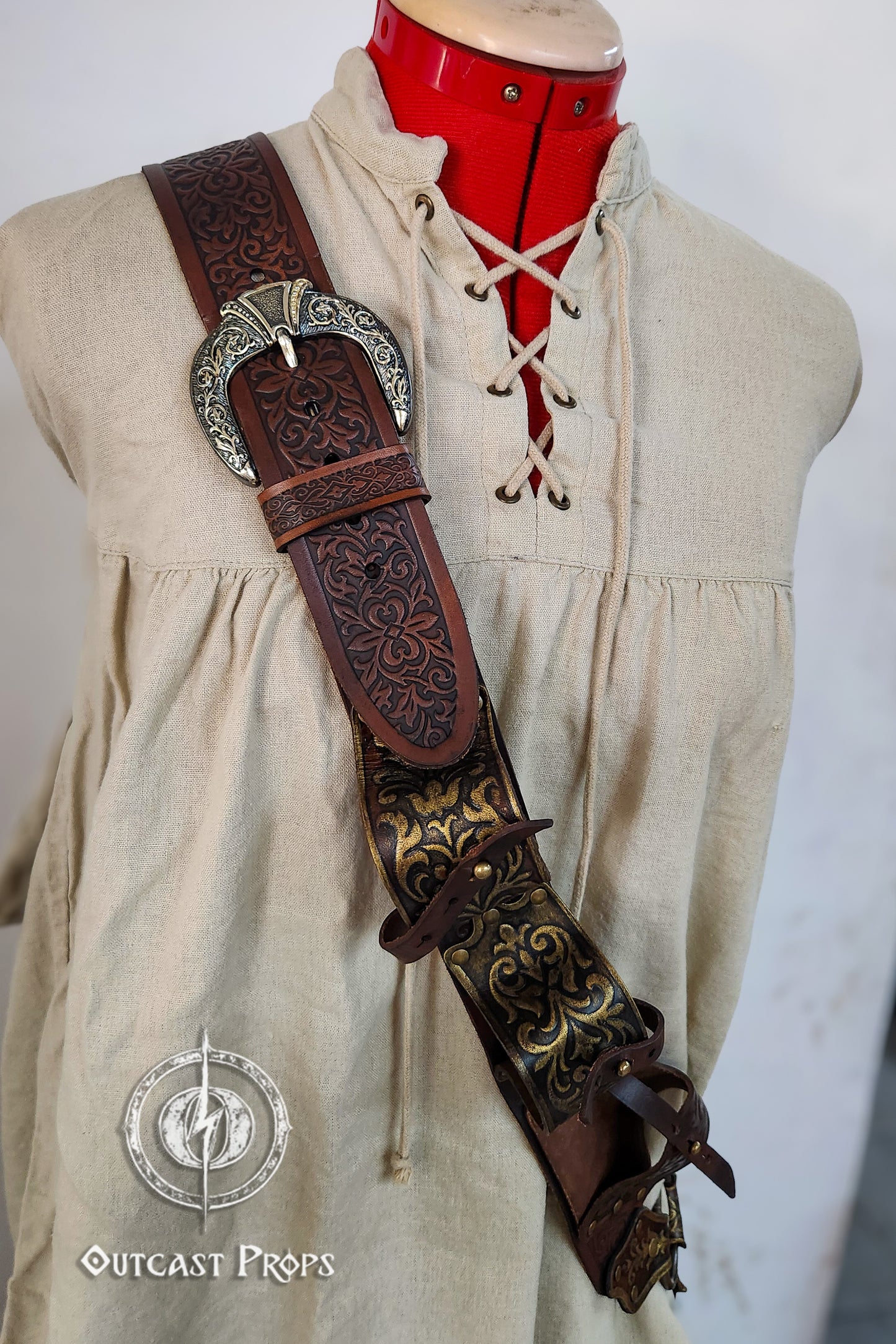 Mannequin wearing a beige dress with a decorative leather belt and sword, branded 'Outcast Props'. Pirate baldric for 4 flintlock pistols and a sword, inspired by Blackbeard, Anne Bonny, Captain Kidd and Pirates of the Caribbean. Ideal for LARP, Renfaire, renaissance fair, Pirate Weekend, cosplay, Halloween and Black Sails. Silver-engraved leather, handmade with vegetable-tanned leather. Premium pirate shoulder belt for Jack Sparrow costume, historical reenactment and fantasy events. High quality pirate acc