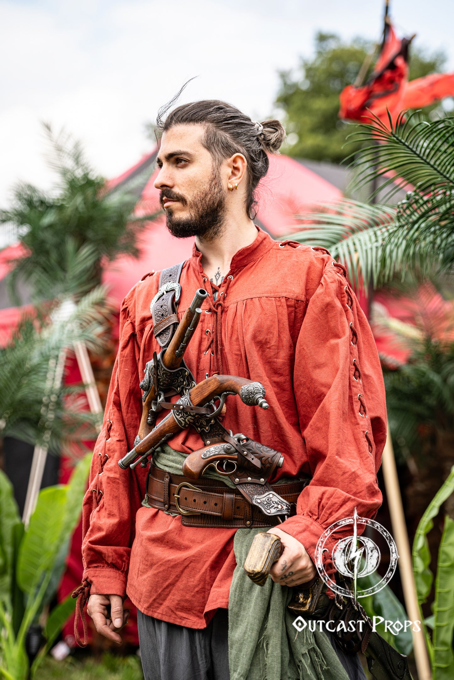 Pirate baldric for 4 flintlock pistols and a sword, worn crossbody over a red shirt. Inspired by Blackbeard and Anne Bonny, this silver-engraved leather shoulder belt is perfect for Jack Sparrow costume, Black Sails fans, LARP, renaissance fair, cosplay or Pirate Weekend. Handmade with vegetable-tanned leather. Premium pirate accessory for reenactment and fantasy events, ideal for themed photoshoots. Man in a red outfit with props, standing outdoors with greenery and tents in the background.