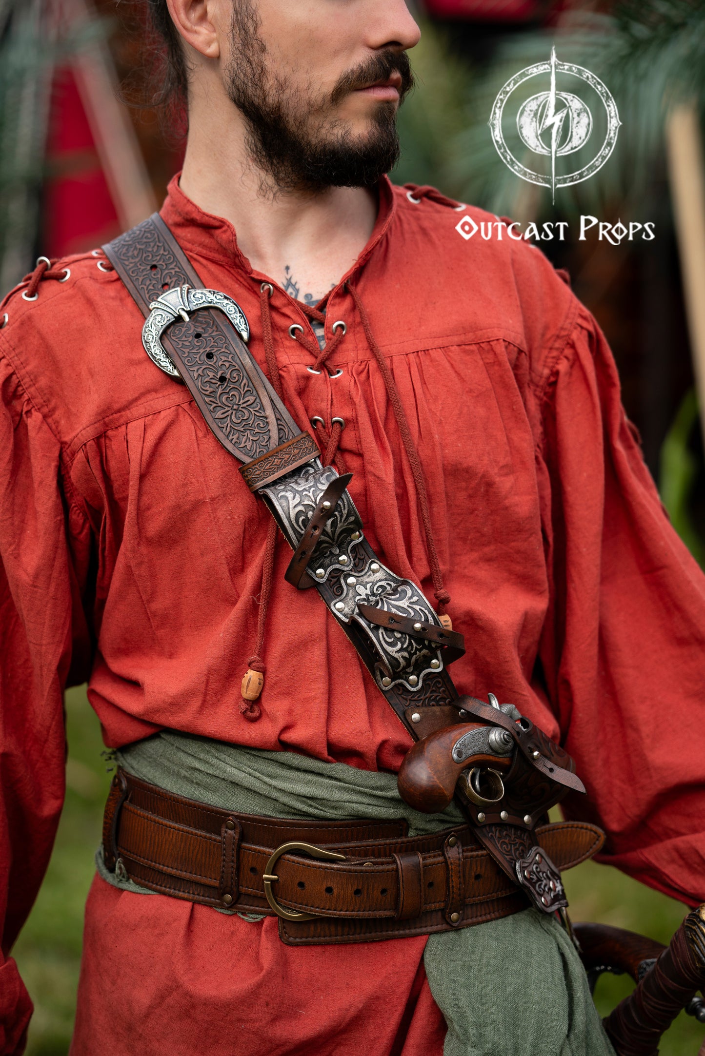 Close-up of pirate wearing silver-engraved leather baldric inspired by Blackbeard and Anne Bonny. Holds flintlock pistols and sword, perfect for LARP, pirate cosplay or Renaissance fairs. Premium handmade belt crafted with vegetable-tanned leather. Ideal for Jack Sparrow costumes, Pirate Weekend and themed events. High detail on buckles, engravings and leather texture. Person wearing a red outfit with a decorative leather strap and Outcast Props logo.