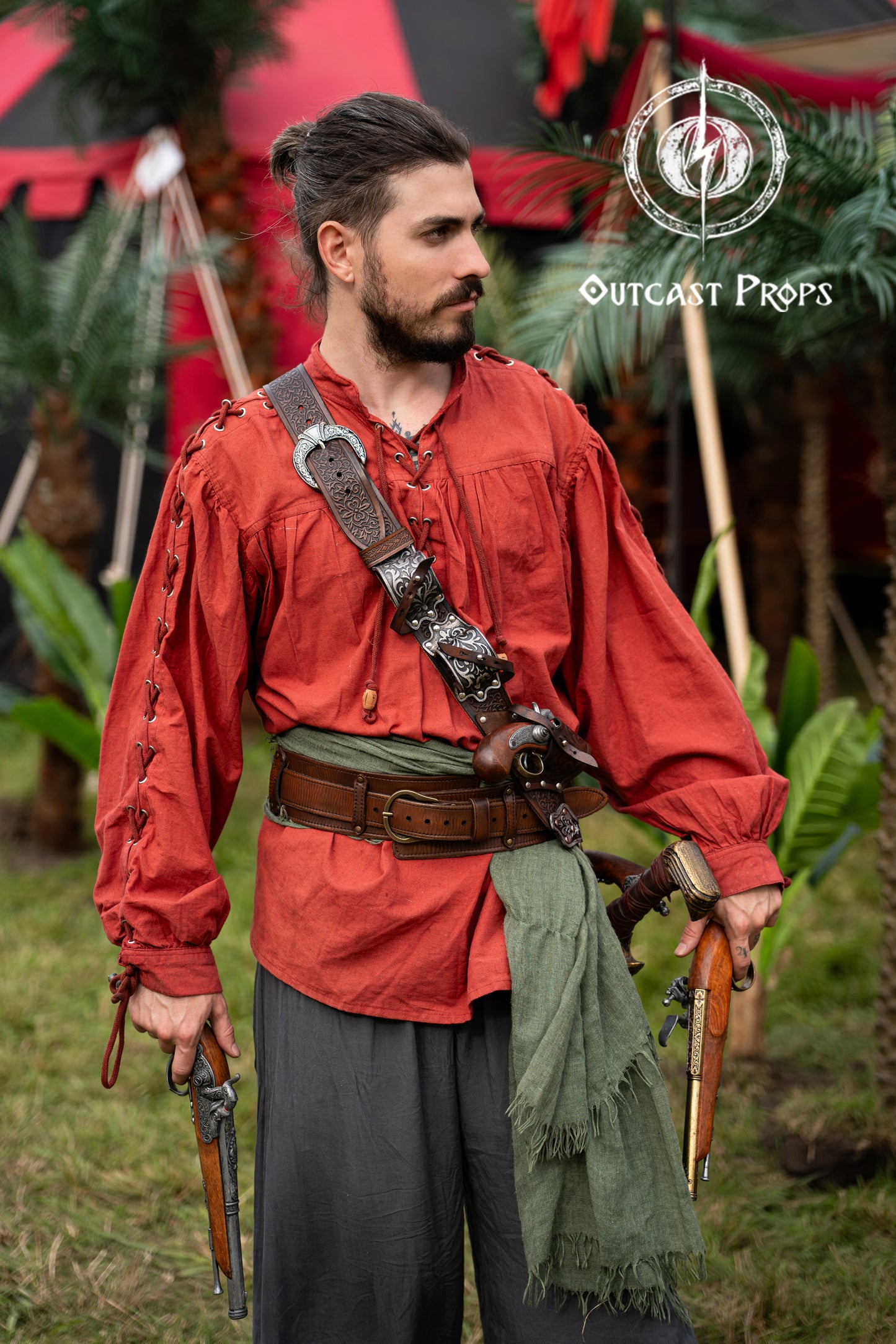 Full pirate outfit with a red shirt, green sash and pirate baldric for 4 flintlock pistols and a sword. Designed for Jack Sparrow fans and pirate reenactors, this silver-engraved leather piece is ideal for LARP, renaissance fairs, cosplay and themed photoshoots. Inspired by Blackbeard and historical pirates, handmade with premium vegetable-tanned leather for an authentic pirate look at any fantasy or Halloween event. Man in historical reenactment attire with weapons and props, standing outdoors.