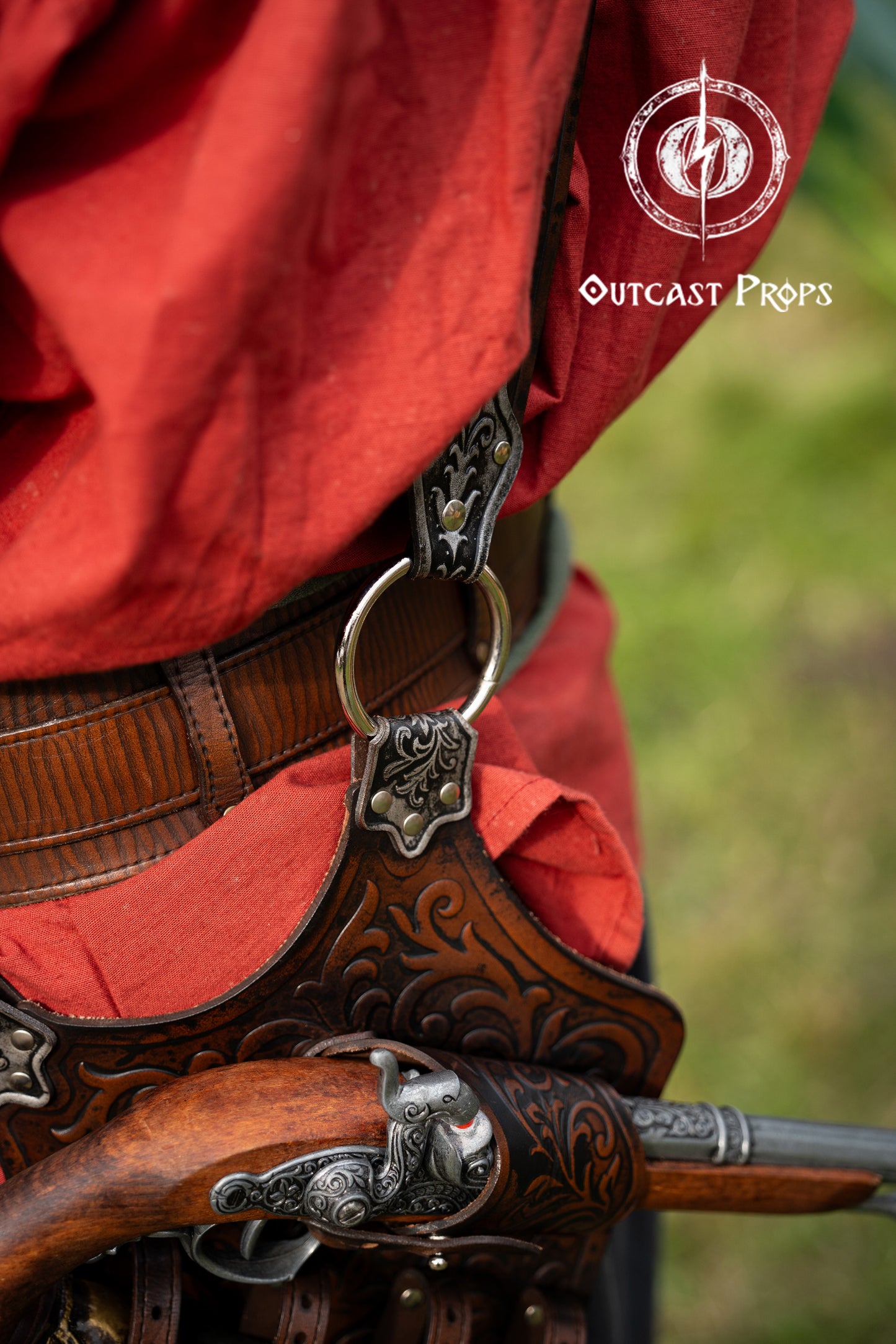 Detailed view of the back connection between the belt and the pirate baldric, showing the metal ring, engraved leather and secured pistol holster. Silver embossing on vegetable-tanned leather, ideal for LARP, Pirate Weekend or Renaissance fairs. Perfect pirate accessory for Blackbeard, Captain Kidd or Jack Sparrow costume. Sturdy and decorative. Close-up of a detailed belt with a sword and red fabric, featuring 'Outcast Props' logo.