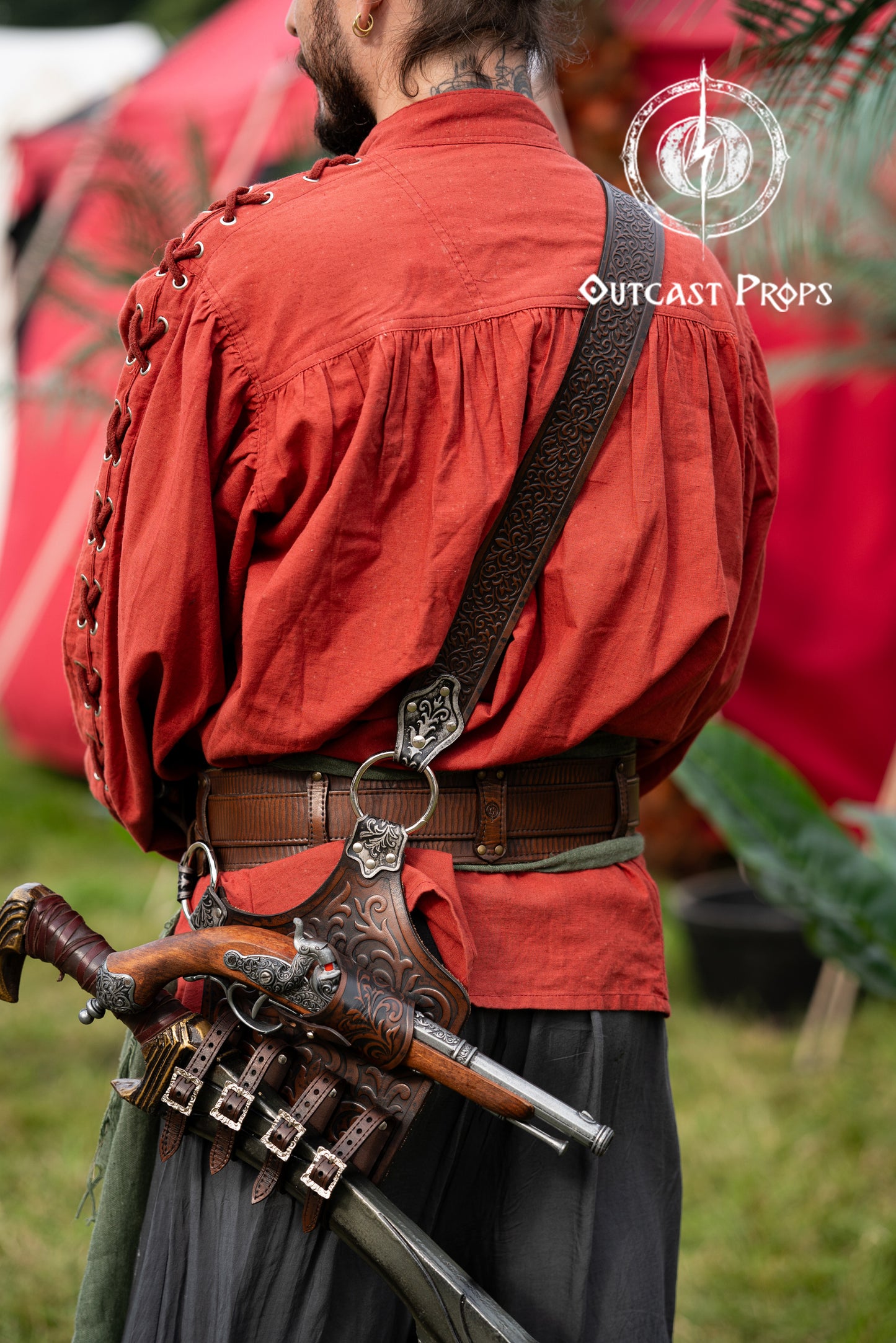 Back view of a pirate wearing the full baldric with four flintlock pistols and a cutlass. Silver engraved leather holsters connected by a wide shoulder strap. Inspired by historical pirates and designed for cosplay, LARP, Pirate Weekend, Halloween and reenactment. Elegant and functional, this baldric is handmade with high-quality vegetable-tanned leather. Person in historical costume with red shirt and gun, standing outdoors with 'Outcast Props' logo.