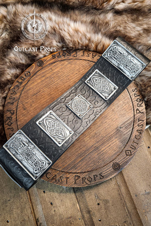 Arabic leather belt with silver plates and floral embossing, displayed over a fur pelt and engraved wooden prop. This wide leather belt is handmade in black vegetable-tanned leather with aged silver details. Perfect for arabic fantasy leather belt, and larp belt for costume. Great for LARP, cosplay, and fantasy characters. Ramattush Persian Leather Broadbelt, ornamental Persian courts inspired, larp oriental leather belt, fantasy belt, renfaire belt. Jafar cosplay costume. Aladdin cosplay.