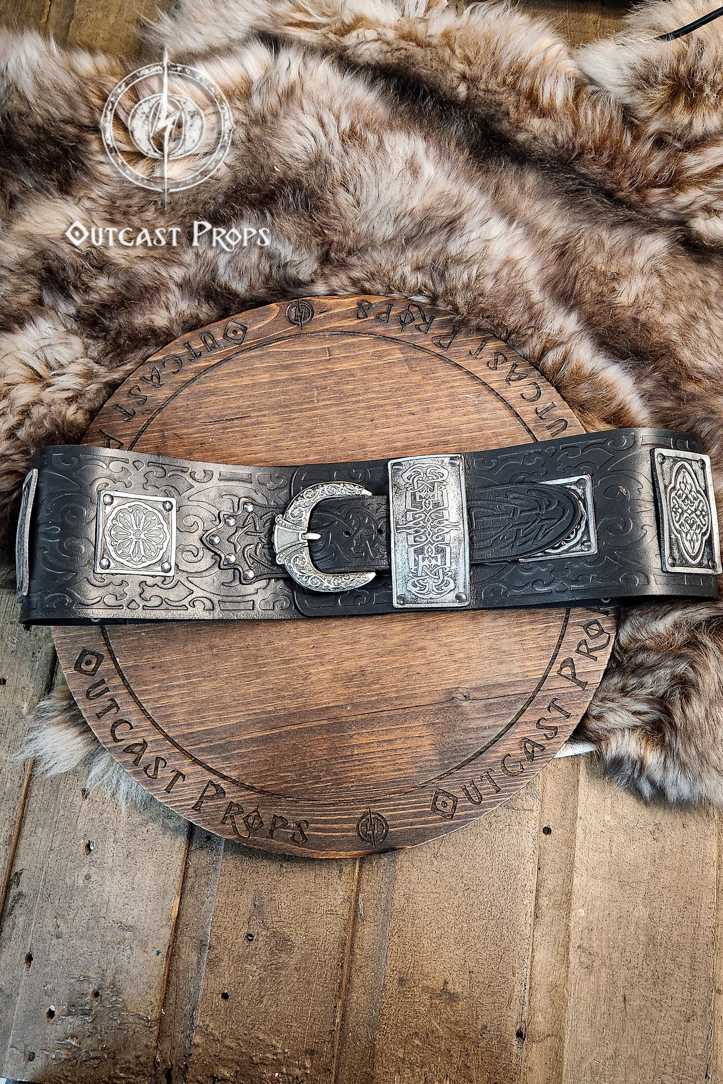 Decorative belt with intricate designs on a wooden surface and fur background, featuring 'Outcast Props' branding. Arabic leather belt with silver plates and floral embossing, displayed over a fur pelt and engraved wooden prop. This wide leather belt is handmade in black vegetable-tanned leather with aged silver details. Perfect for arabic leather belt, fantasy leather belt, and larp belt for costume. Great for LARP, cosplay, and fantasy characters.