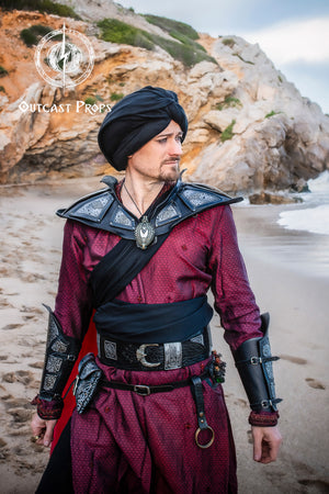 Person in medieval or fantasy costume standing on a beach with rocky cliffs in the background. Ramattush Persian Leather Broadbelt, ornamental Arabic and Persian courts inspired, larp oriental leather belt, fantasy belt, renfaire belt. Jafar cosplay costume. Aladdin cosplay.