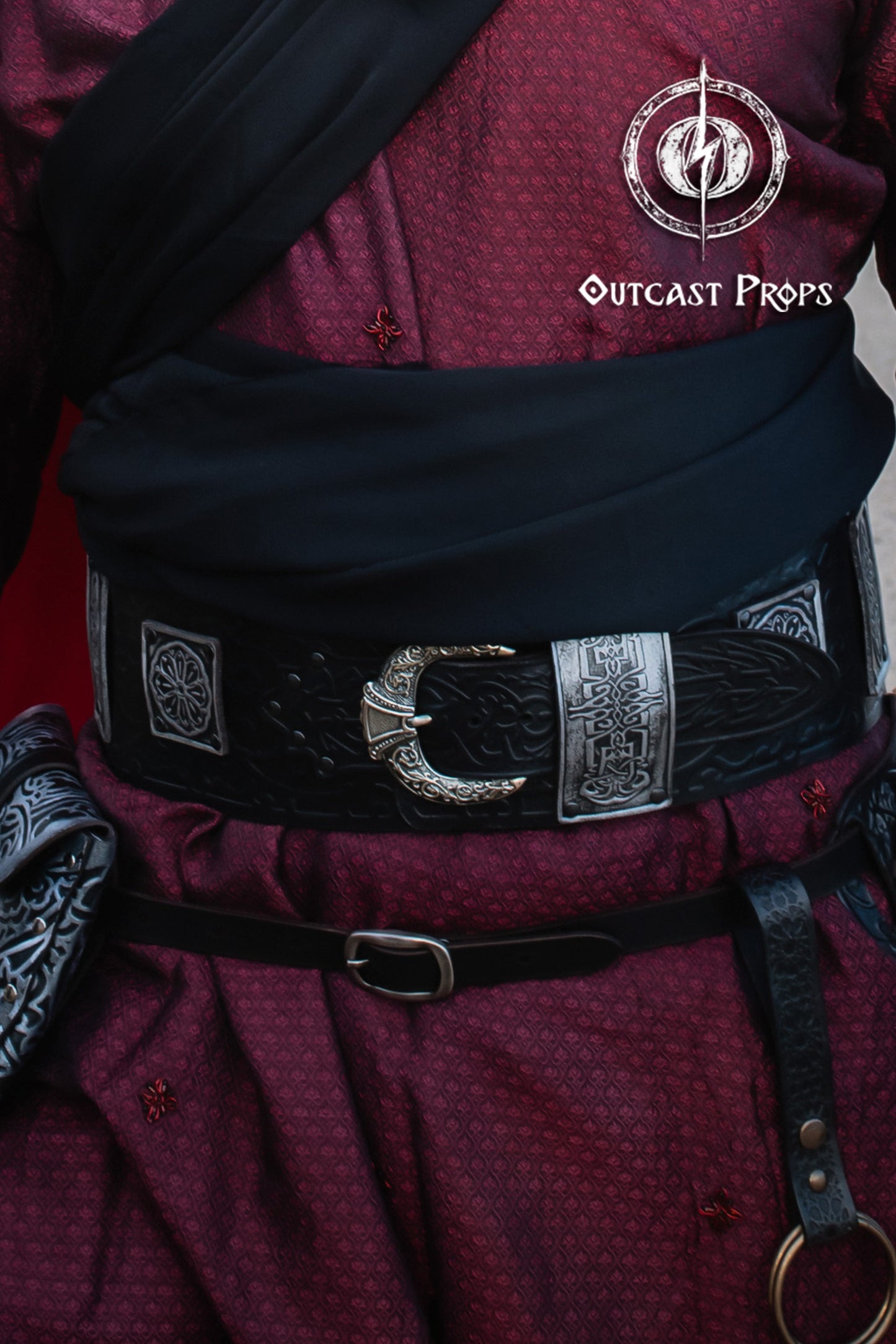 Person wearing a detailed belt with decorative buckles and 'Outcast Props' logo. Close-up of wide arabic leather belt worn over red robes. Shows the craftsmanship of silver elements and embossed black leather. Perfect SEO alt text for wide leather belt, fantasy leather belt, and leather belt for costume.