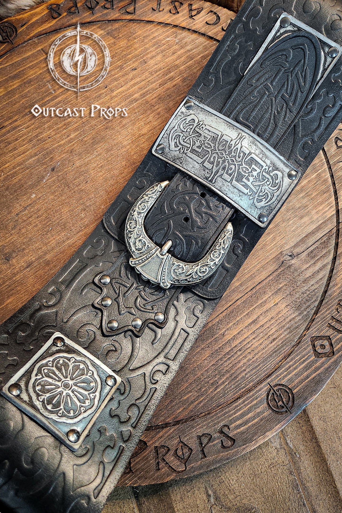 Decorative belt with intricate designs on a wooden surface, featuring the brand 'Outcast Props'. Detailed view of crescent-shaped silver buckle and engraved plates on black leather. This persian leather belt combines historical elegance and sturdy design. Crafted for LARP belt for costume and fantasy outfits.