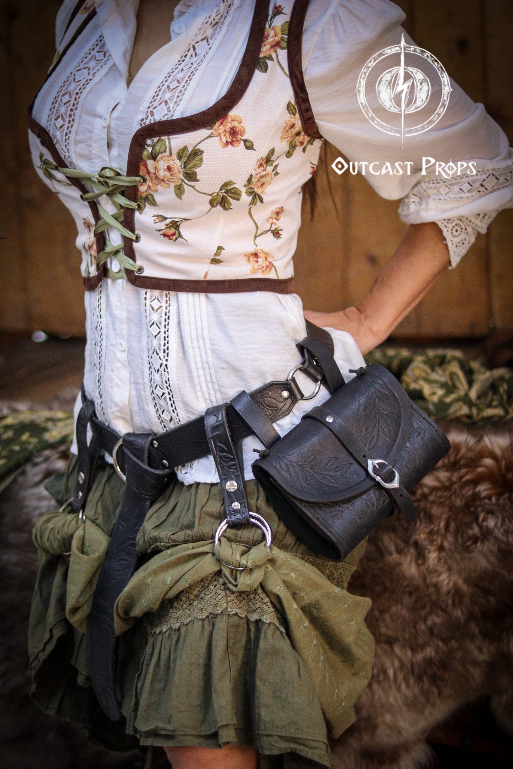 Cottagecore and fantasy-inspired outfit with a black floral tooled leather belt set, worn over a white lace blouse and green gathered skirt. Includes hip bag and skirt hikes for Ren Faire, LARP, or medieval cosplay events. Handmade from Spanish full-grain leather, dyed with natural pigments, combining rustic charm with practical utility for adventurers.