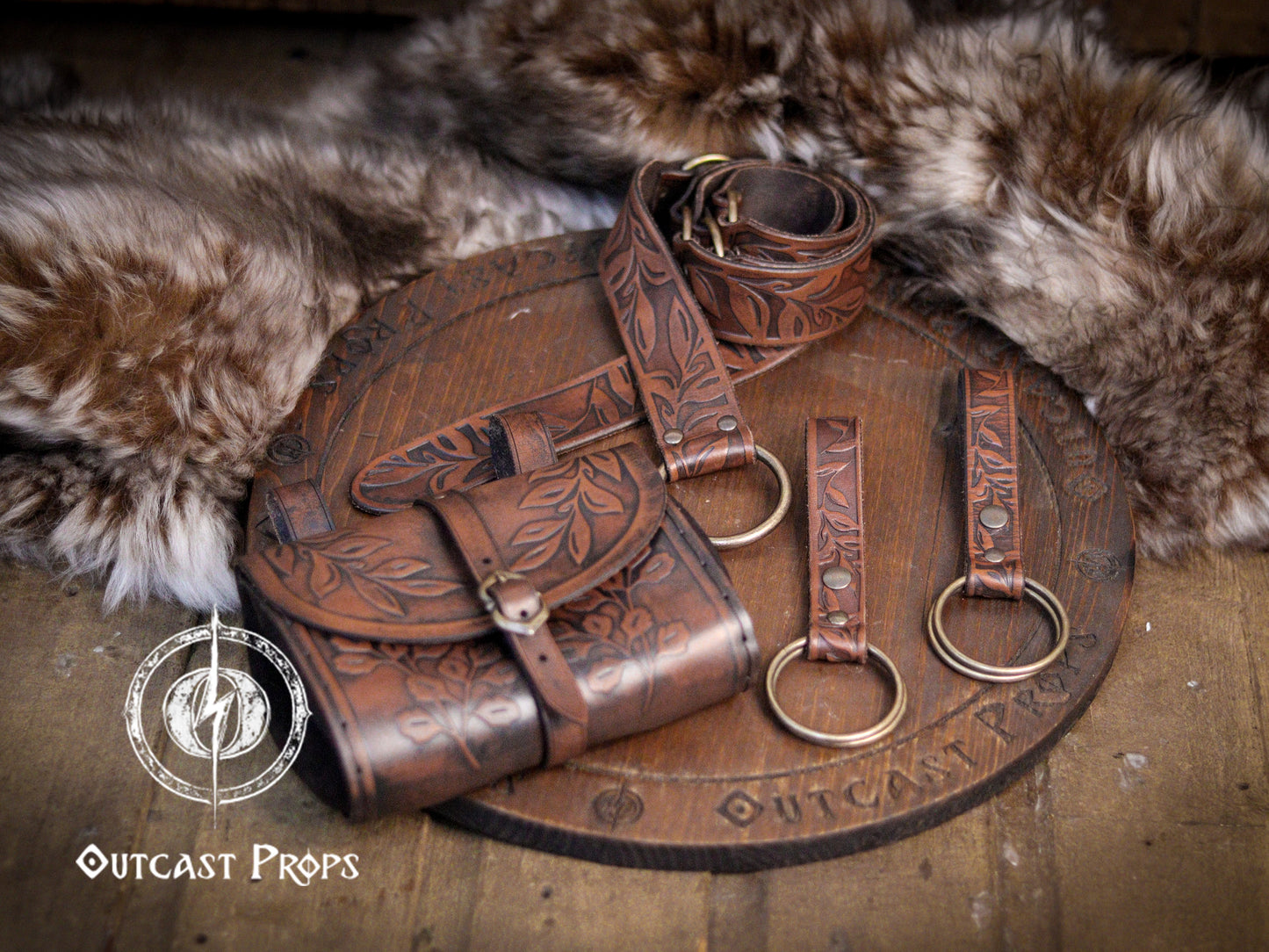 Full fantasy leather set with floral embossing, including belt with O-rings, hip bag, and skirt hikes, crafted by Outcast Props. Shown on a wooden board surrounded by natural furs, evoking medieval and fantasy aesthetics. Ideal for Ren Faires, LARP, cosplay, pirate, or witchy costumes. Made of vegetable-tanned leather, hand-tooled and dyed for a unique, story-rich appearance.