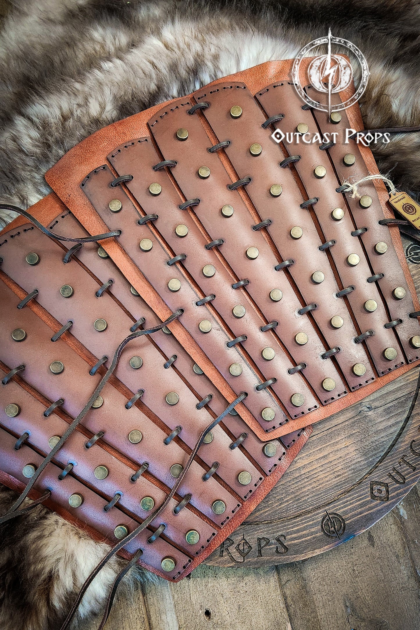 Brown leather armor with metal studs on a wooden surface, branded 'Outcast Props'. Samurai Leather Greaves for LARP Cosplay or Renaissance Fair Studded Shin Armor Fantasy Warrior Leg Guards Handmade Medieval Costume Piece