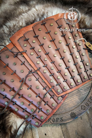 Brown leather armor with metal studs on a wooden surface, branded 'Outcast Props'. Samurai Leather Greaves for LARP Cosplay or Renaissance Fair Studded Shin Armor Fantasy Warrior Leg Guards Handmade Medieval Costume Piece