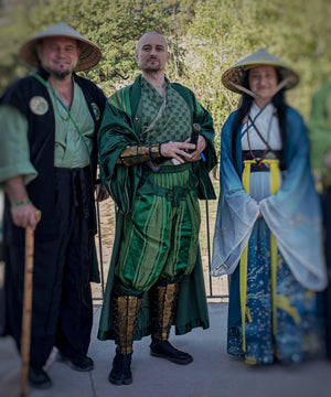 Full view of a person in green samurai costume standing between two others, wearing golden dragon shin guards and bracers over traditional layered robes. The outfit is styled for Japanese-inspired cosplay or fantasy LARP character portrayal.